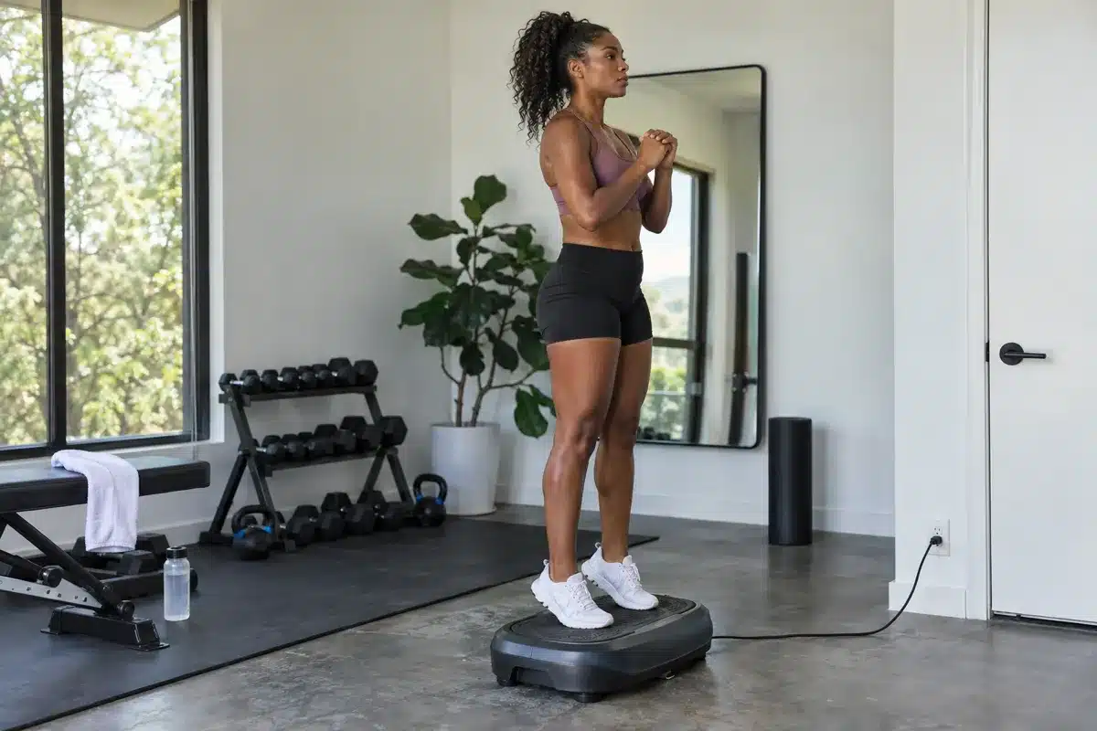 An African American woman standing in a relaxed warm-up stance on a gray vibration plate in a modern home gym.
